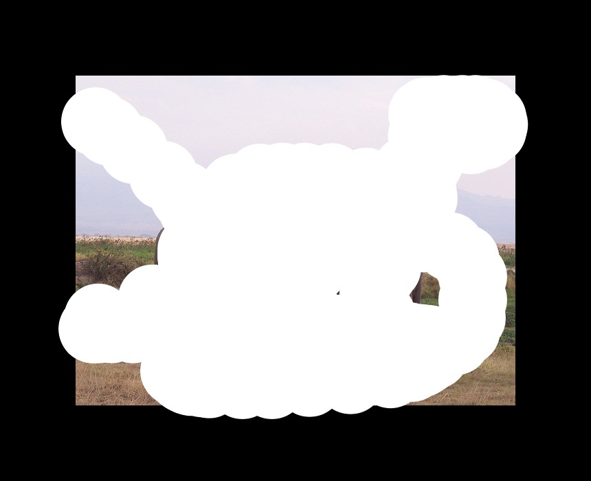 In the center of the image there are two elephants. At the bottom there is grass. In the background we can see hills and sky.