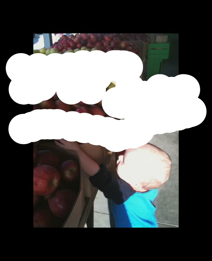 In this picture we can see a baby standing near to the apple boxes and touching the apples, apples are green, and red in color.